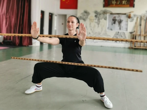 Student in Ma Bu stance balancing two sticks—one across the thighs and one extended in the arm.